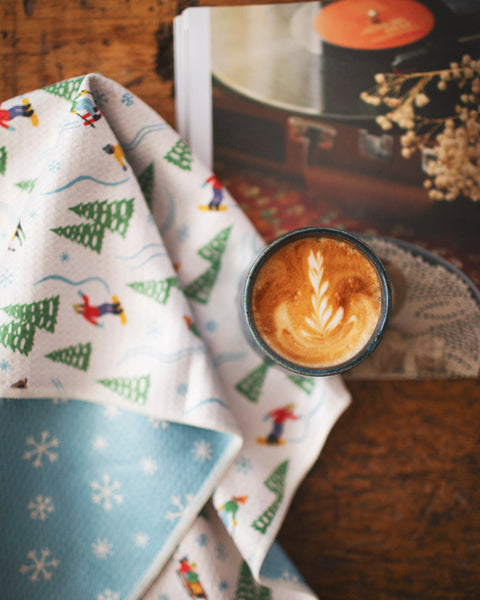 Snow Day Kitchen & Tea Towel displayed with a latte and a magazine, showcasing winter vibes and ski design.