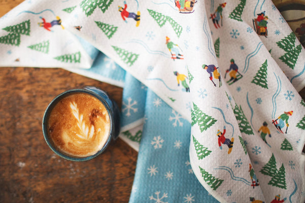 Snow Day Kitchen & Tea Towel with vibrant winter design, eco-friendly, super absorbent Grip-Tex™ fabric, displayed with a cup of coffee.