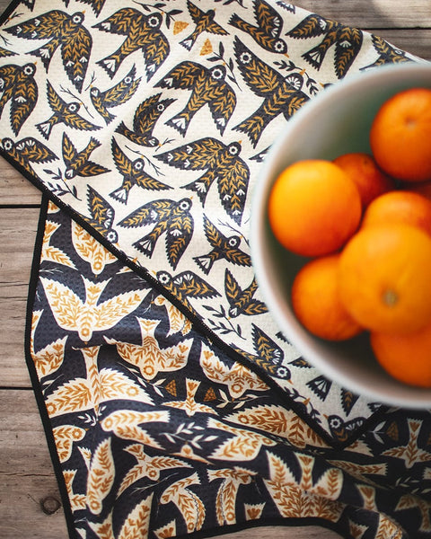 Songbird Kitchen & Tea Towel with bird pattern next to a bowl of oranges on a wooden counter, eco-friendly and super absorbent.