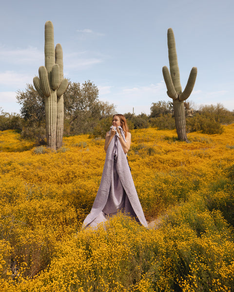 Woman wrapped in a lavender handwoven Sonoma Blanket, standing in a cactus-filled desert landscape, exuding beauty and coziness. By Trek Light.