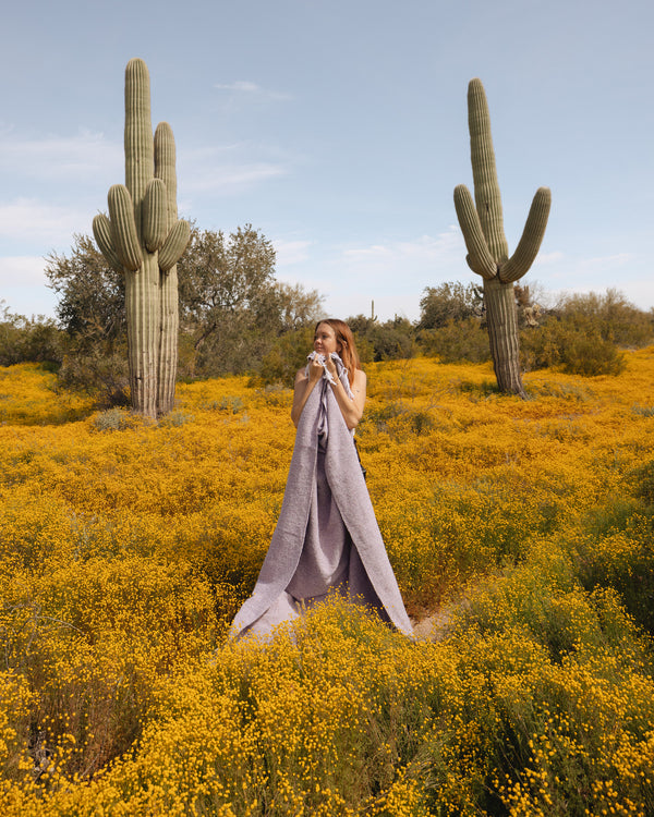 Woman wrapped in a lavender handwoven Sonoma Blanket, standing in a cactus-filled desert landscape, exuding beauty and coziness. By Trek Light.