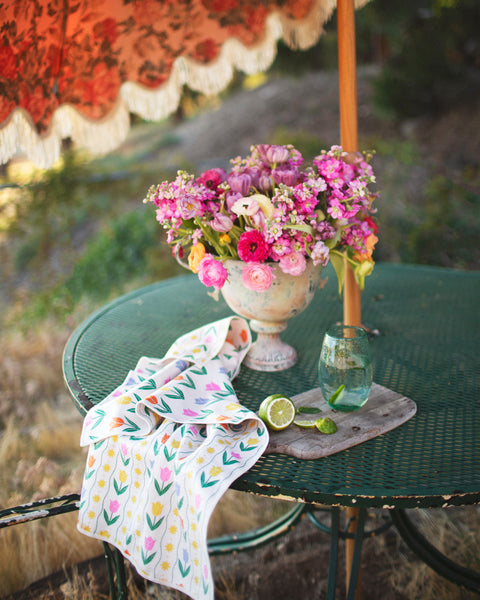 Spring Tulips Kitchen & Tea Towel draped on a green table, with a flower vase and refreshing drink.