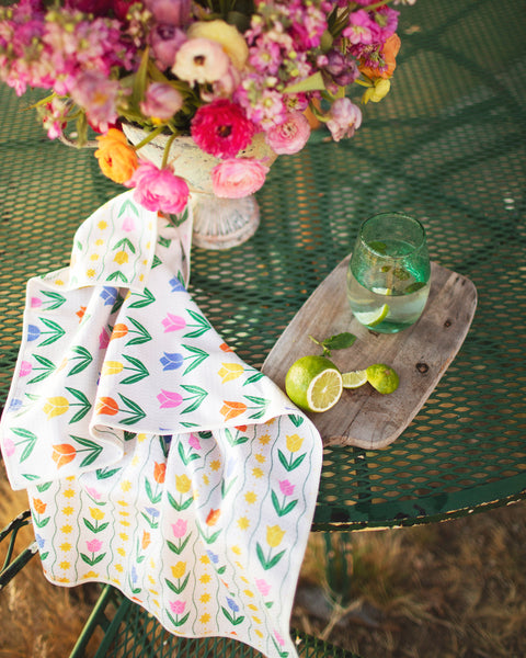 Spring Tulips Kitchen & Tea Towel draped on a green table with flowers and drink, showcasing its colorful floral design.