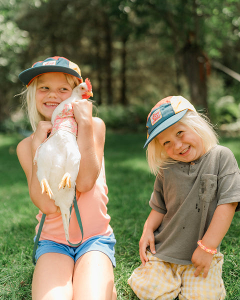 Kids wearing Stay Wild Kids Hat while holding a chicken outdoors in a playful summer setting.