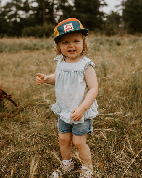 Child wearing colorful Trek Light Stay Wild hat, smiling in a field, ready for outdoor adventure. Perfect 5-panel camper style for kids.
