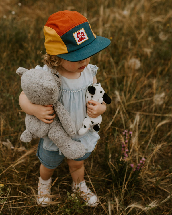 Child wearing colorful Trek Light Stay Wild hat, holding stuffed animals in a grassy field, ready for adventure.