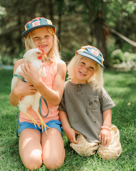 Children wearing Stay Wild Kids Hat while holding a chicken in a sunny outdoor setting.