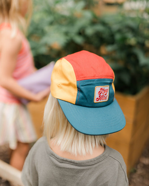 Stay Wild Kids Hat displayed on a child in a playground setting, featuring a colorful 5-panel design.