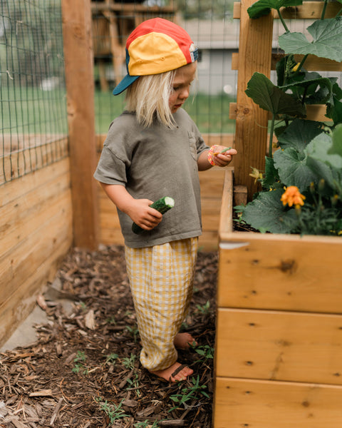 Child wearing Stay Wild Kids Hat while exploring a garden, holding a cucumber and enjoying outdoor play.