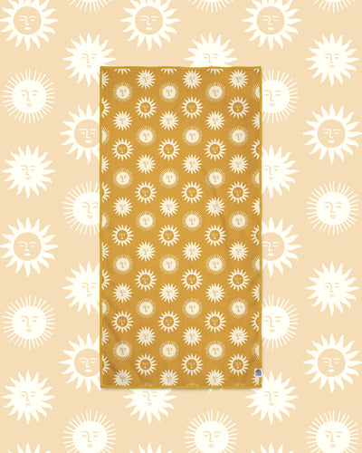 Sunny Wander & Travel Towel in mustard yellow with cream sun face print, laid flat on a peach background of larger suns.