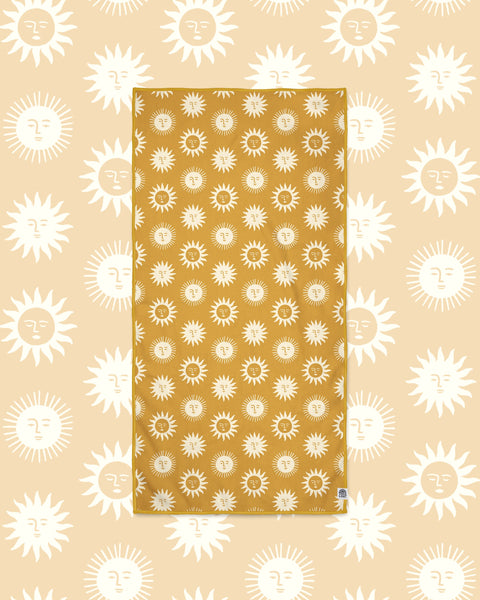 Sunny Wander & Travel Towel in mustard yellow with cream sun face print, laid flat on a peach background of larger suns.