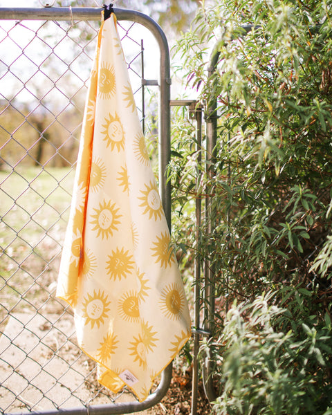 Sunny Wander & Travel Towel hanging on a fence, showcasing its bright sunshine design.