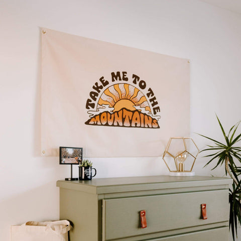 Take Me To The Mountains Canvas Flag with Trek Light design, natural canvas, and easy hanging brass grommets, displayed indoors.