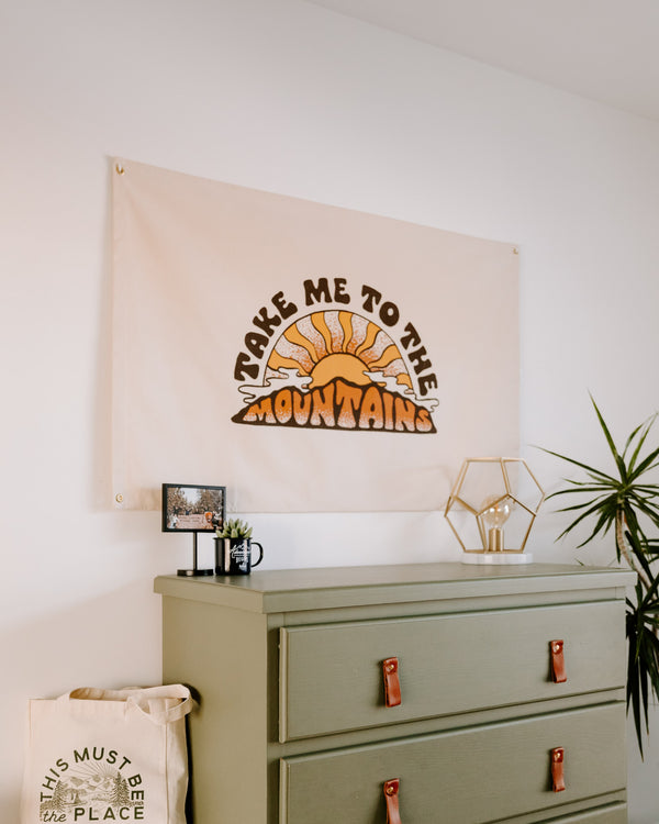 Take Me To The Mountains Canvas Flag, cream with a retro sun and mountain print, hung above a green dresser.