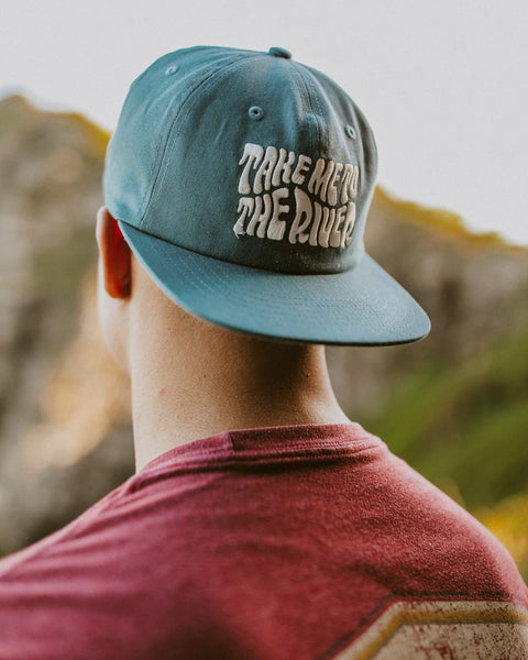 Take Me To The River Hat in teal with white wavy embroidered text, worn by a man in a red shirt facing blurred cliffs.