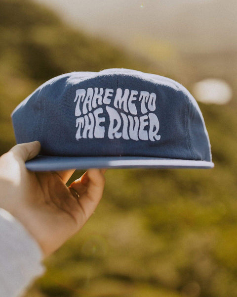 A hand holds the Take Me To The River Hat, a faded blue cap with white wavy lettering, against a blurred green hillside.