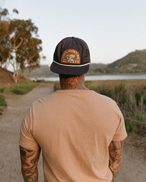 Person wearing This Must Be The Place Hat (Mountains) while walking on a scenic pathway near water.