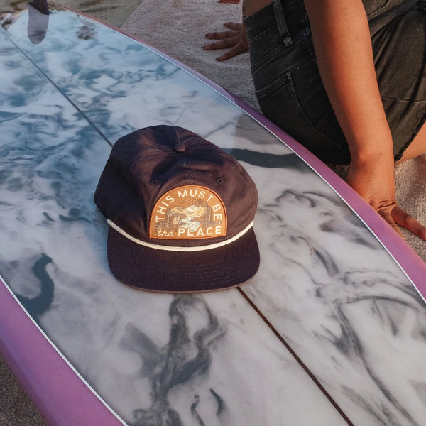 This Must Be The Place Hat (Mountains) resting on a surfboard at sunset, showcasing its stylish design and comfort.