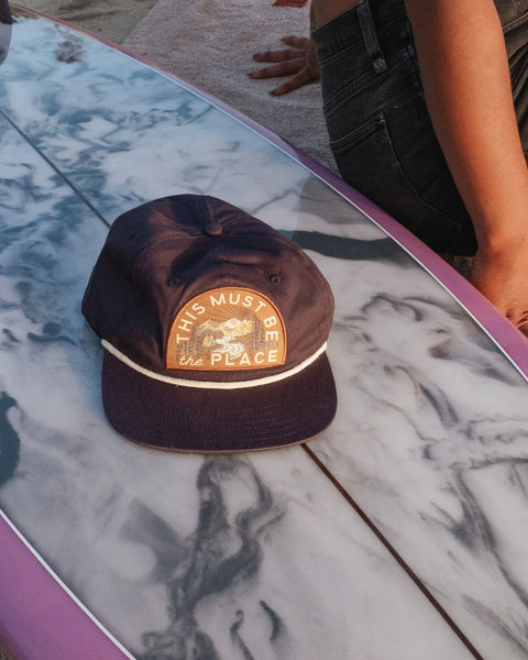 This Must Be The Place Hat (Mountains) resting on a surfboard at sunset, showcasing its stylish design and comfort.