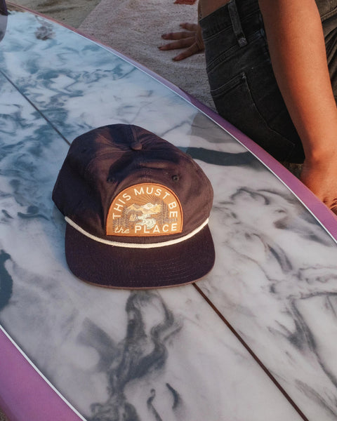 This Must Be The Place Hat in Mountains, navy with a rope trim and patch, on a marbled surfboard beside a seated person.