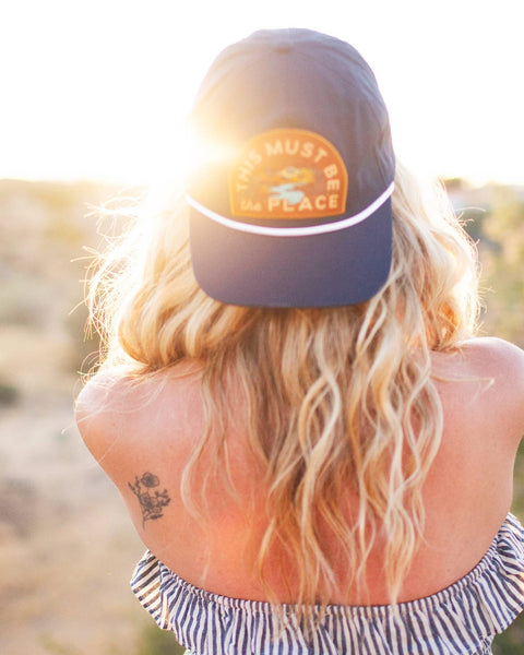 Woman wearing This Must Be The Place Hat (Mountains) with sun flare in the background, ideal for camping and outdoor adventures.