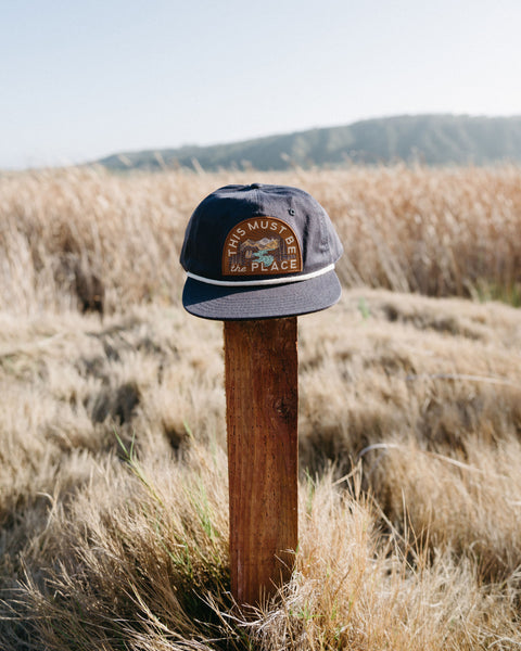 This Must Be The Place Hat (Mountains) displayed on a wooden post in a grassy field with mountains in the background.