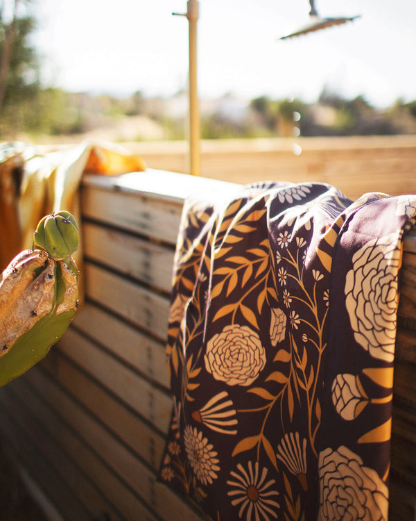 alt_tag: Travelin' Light Wander & Travel Towel hanging by an outdoor shower, showcasing its vibrant floral design.