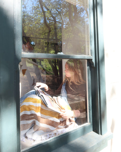 Woman reading while wrapped in Golden Age Blanket by a window, enjoying comfort and the view outside.