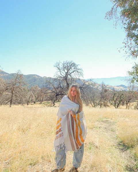 Golden Age Blanket draped over a woman in a field under blue skies, perfect for cozy outdoor moments.