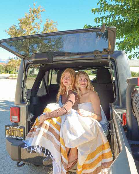 Two women wrapped in Golden Age Blankets sitting in the back of a vehicle, enjoying a sunny day outdoors.