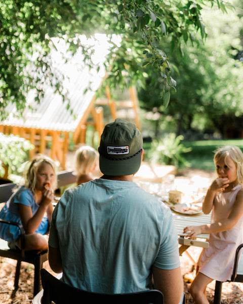 A person wearing a Trout Hat (Olive) enjoys a meal outdoors with children, showcasing a relaxed lifestyle.