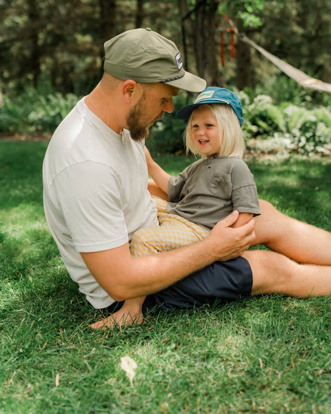 Dad in an olive Trout Hat and a child in a blue Trout Hat sitting together on backyard grass, Trek Light