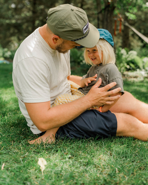 Man in an olive Trout Hat leaning toward a child in a blue Trout Hat on a green lawn, Trek Light
