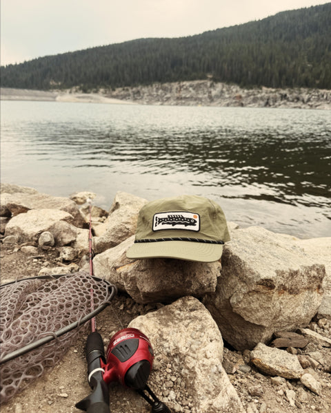 Trout Hat (Olive) resting on rocks by the water, perfect for fishing and outdoor adventures.