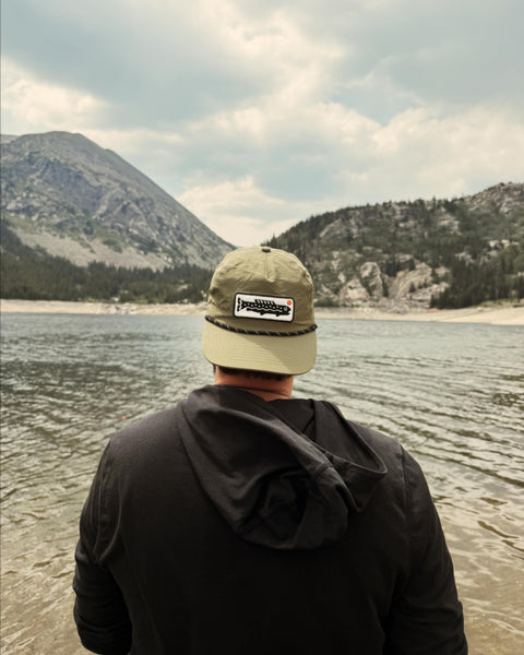 Person wearing a Trout Hat (Olive) by a lake with mountains in the background.