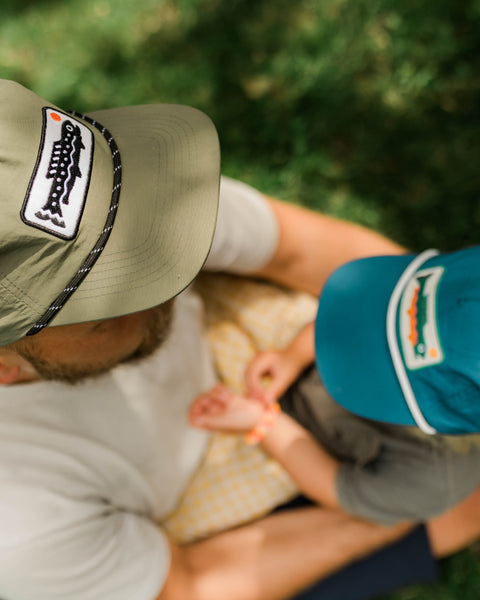 Overhead view of an adult wearing the olive Trout Hat with a fish patch, beside a child in a teal cap, on grass.