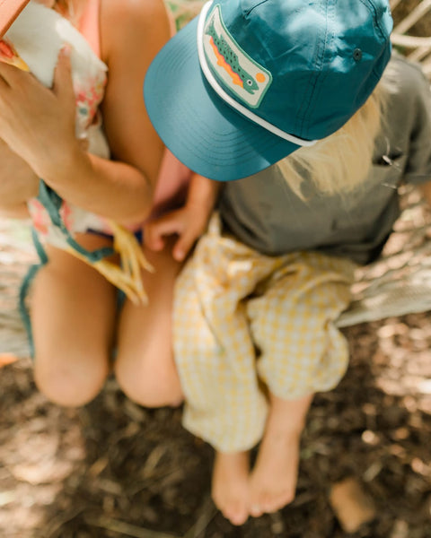 Toddler wearing a Trout Kids Hat while sitting on a hammock with a sibling, enjoying outdoor fun.