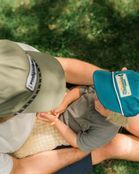 Trout Kids Hat being worn by a child sitting with an adult outdoors on grass.