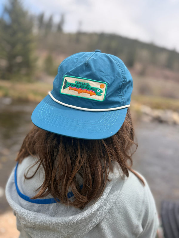 Child wearing a blue Trout Hat Twinsie Set by a river with trees in the background.