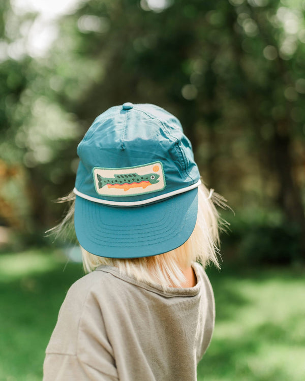 Child in a blue nylon Trout Hat, Twinsie Set Blue, with a fish patch and white rope trim, outdoors by trees.