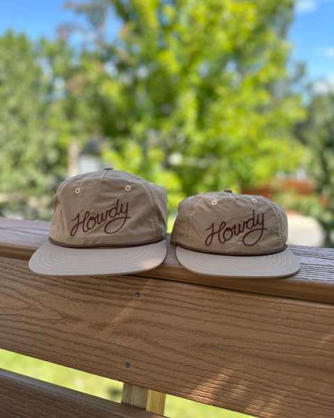 Howdy Hat Twinsie Set of two tan nylon caps with maroon script embroidery resting on a wooden deck rail with trees behind.