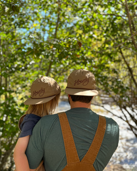 Howdy Hat Twinsie Set in tan with brown script, worn by an adult and child seen from behind in the woods