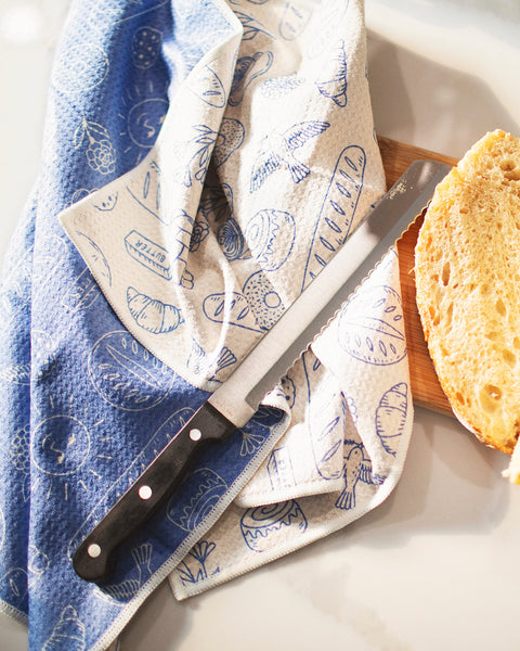 Wake & Bakery Kitchen & Tea Towel with a loaf of bread and a knife on a wooden cutting board.