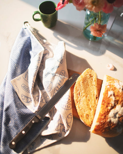 Wake & Bakery Kitchen & Tea Towel on a table with sliced bread, knife, and flowers in a vase.