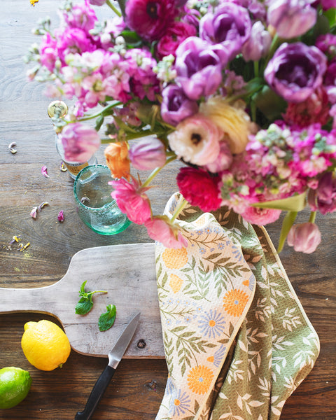 Wavy Flowers Kitchen & Tea Towel displayed on a wooden table with fresh lemons, limes, and a floral arrangement.