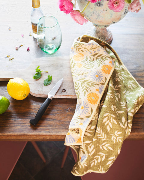 Wavy Flowers Kitchen & Tea Towel on a wooden table with fruits, flowers, and a glass, showcasing its floral design.