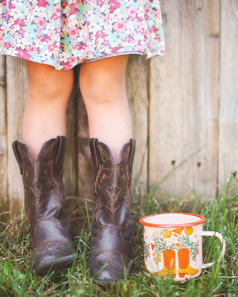 Western Bloom Enamel Mug 16oz by Trek Light with cowboy boots and floral dress in a rustic outdoor setting.