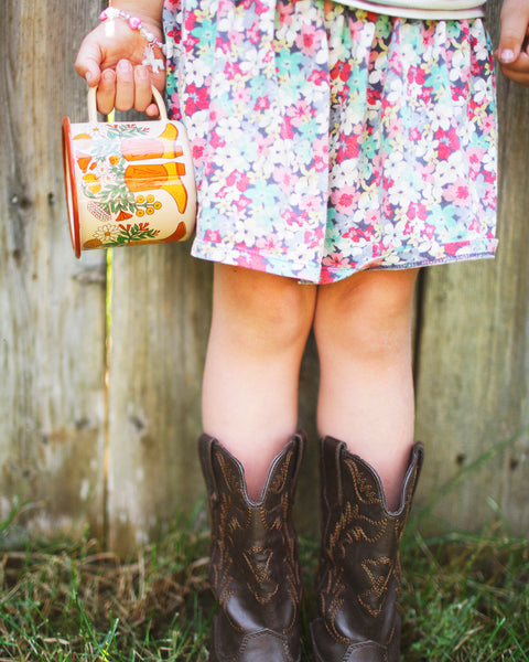 Western Bloom Enamel Mug 16oz in a child's hand, paired with brown cowboy boots and a floral dress.