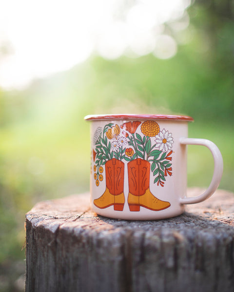 Western Bloom Enamel Mug 16oz with floral and cowboy boot design on a wooden surface outdoors.