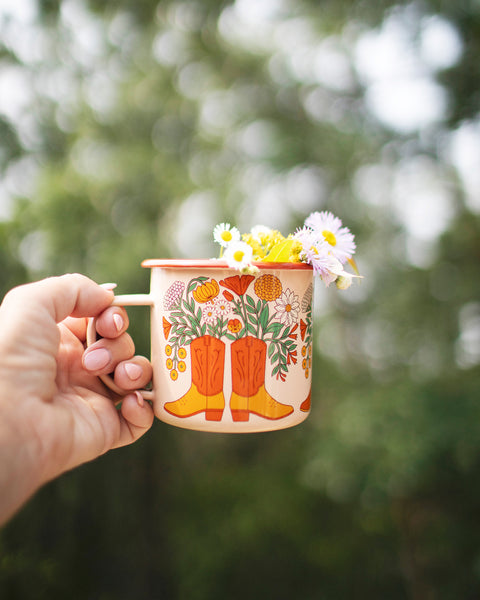 Western Bloom Enamel Mug 16oz with floral design and country boots, held outdoors with soft background blur.